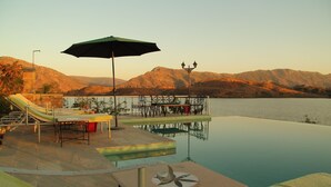 An infinity pool with a view of mountains, lounge chairs, and an umbrella. - Jhadol Safari Resort (Jharol)