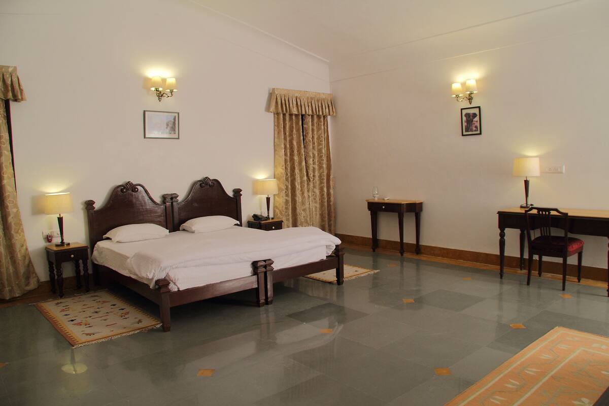 Um quarto bem arrumado com uma cama de casal, mesas de cabeceira com abajures, uma escrivaninha com cadeira e um tapete estampado no chão.