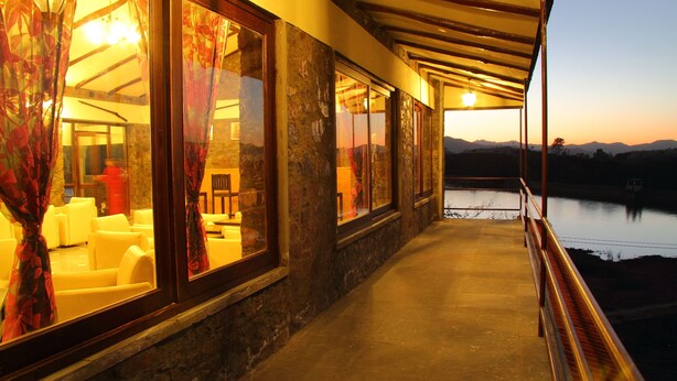 A covered balcony with a view of a calm body of water and distant mountains. - Jhadol Safari Resort (Jharol)