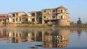 A waterfront property with multiple buildings, including a stone structure with a red roof and a modern building with balconies. - Jhadol Safari Resort (Jharol)