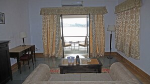 A living room with a sofa, a coffee table, a desk, and a chair. There is a view of a balcony through a glass door. - Jhadol Safari Resort (Jharol)