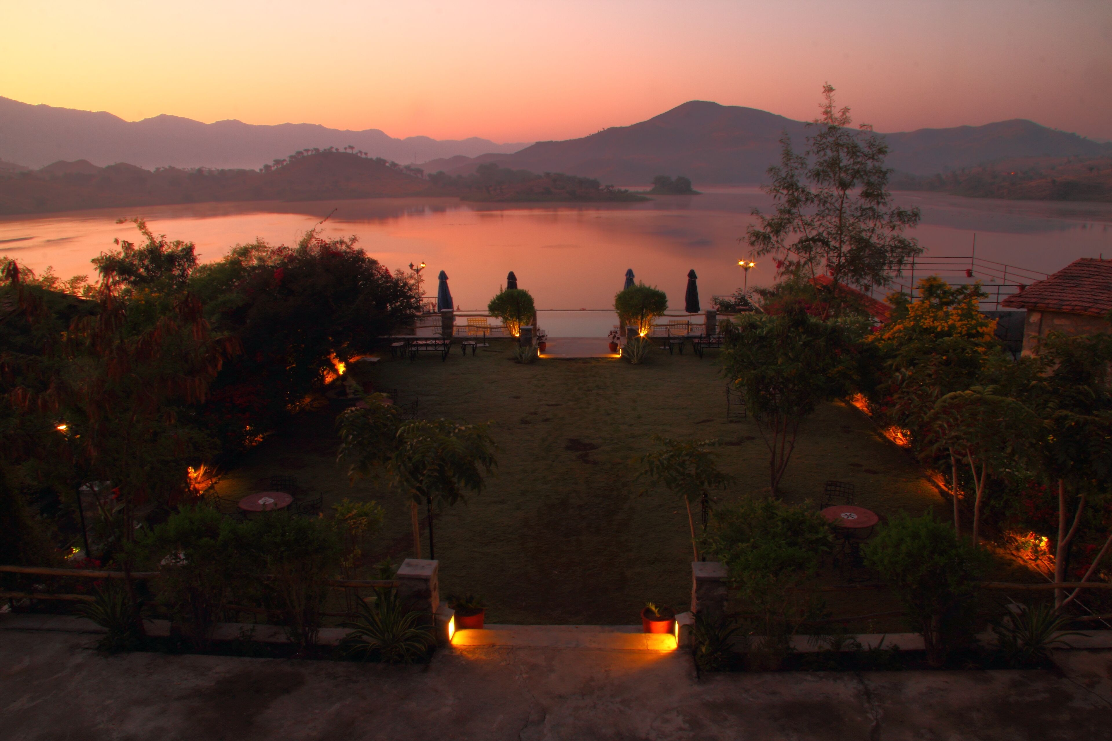 A serene lakeside view at dusk with a patio area featuring tables and chairs, surrounded by lush greenery and a distant mountain range.