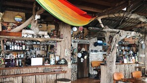 Un bar rústico con estantes de madera, un mostrador de madera, y una variedad de botellas y vasos. Hay sillas y una mesa para sentarse.