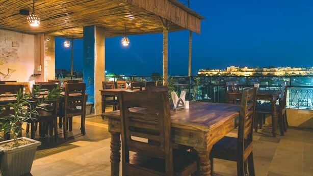 An outdoor dining area with wooden tables and chairs, a covered seating area, and a view of the city at night. - Prithvi Palace (Jaisalmer)
