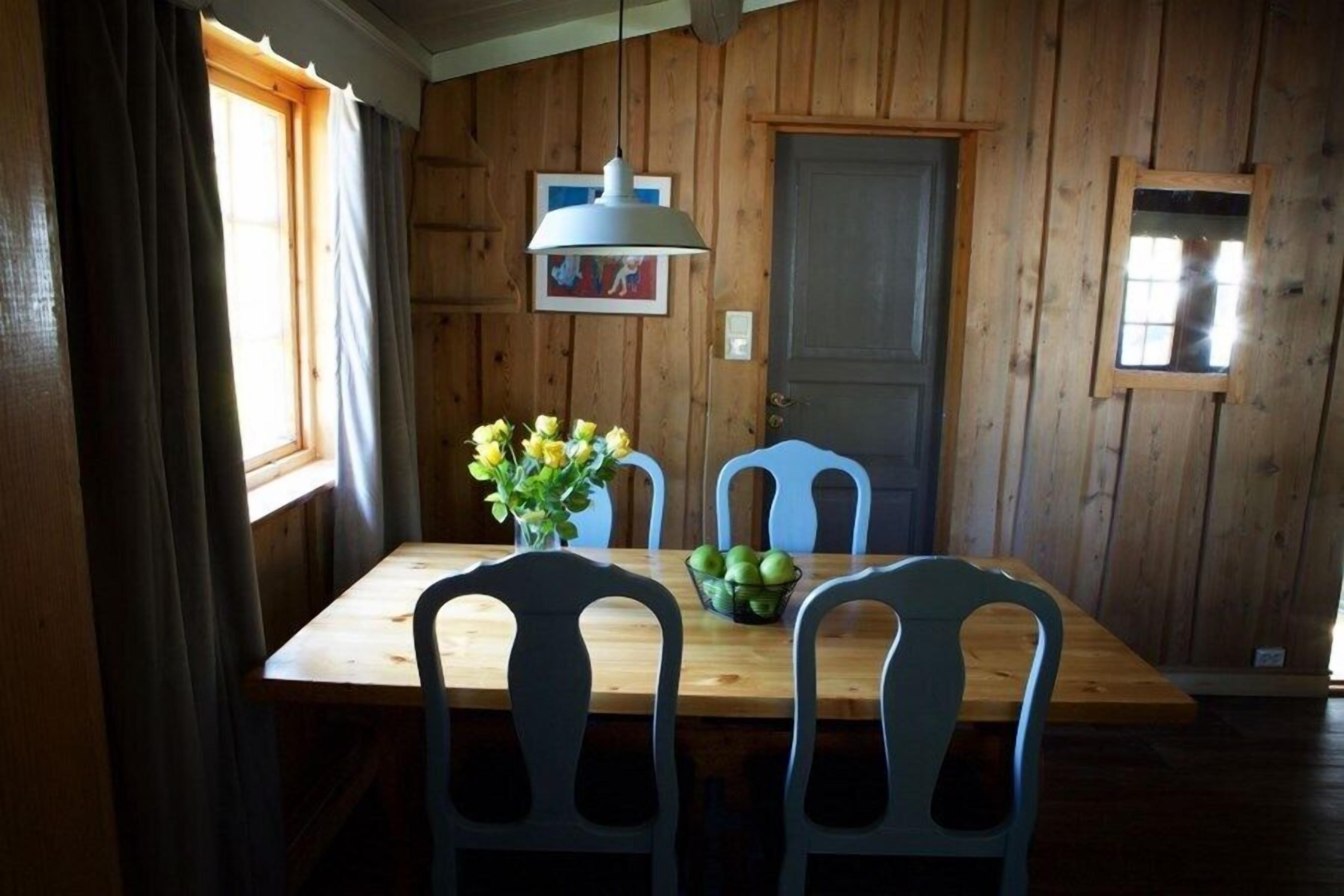 A wooden-paneled room with a wooden table and four chairs. There is a window with curtains, a hanging light fixture, and a door.