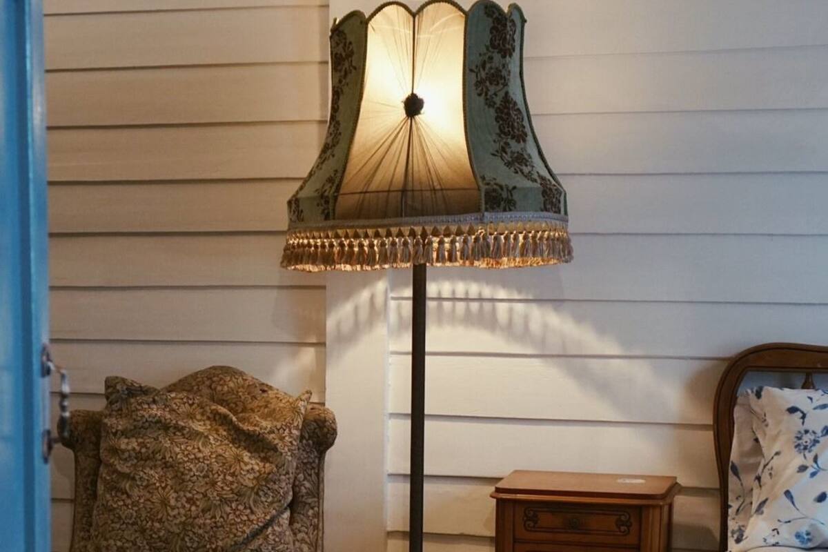 Ett rum med en mönstrad fåtölj, en golvlampa, ett träbord som används som sängbord och en säng med blommig sängklädsel.