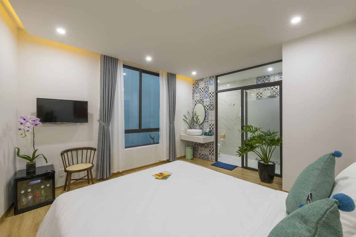 A modern hotel room with a large bed, a flat-screen TV, a wooden chair, a small table with a plant, and a sliding glass door leading to a balcony.