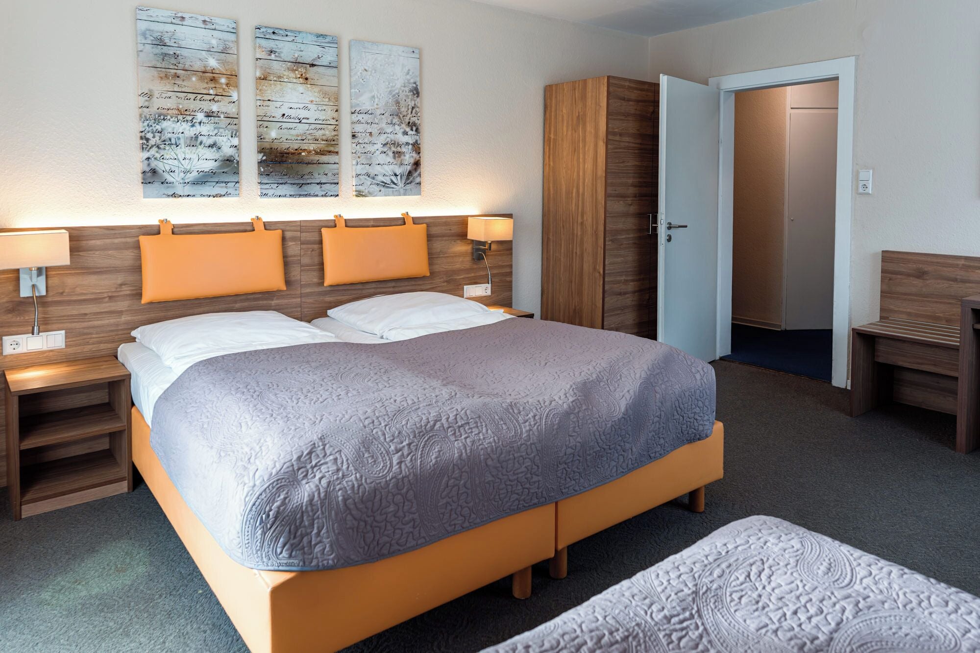 A modern hotel room with a large bed, two bedside tables with lamps, a wardrobe, and a bench.