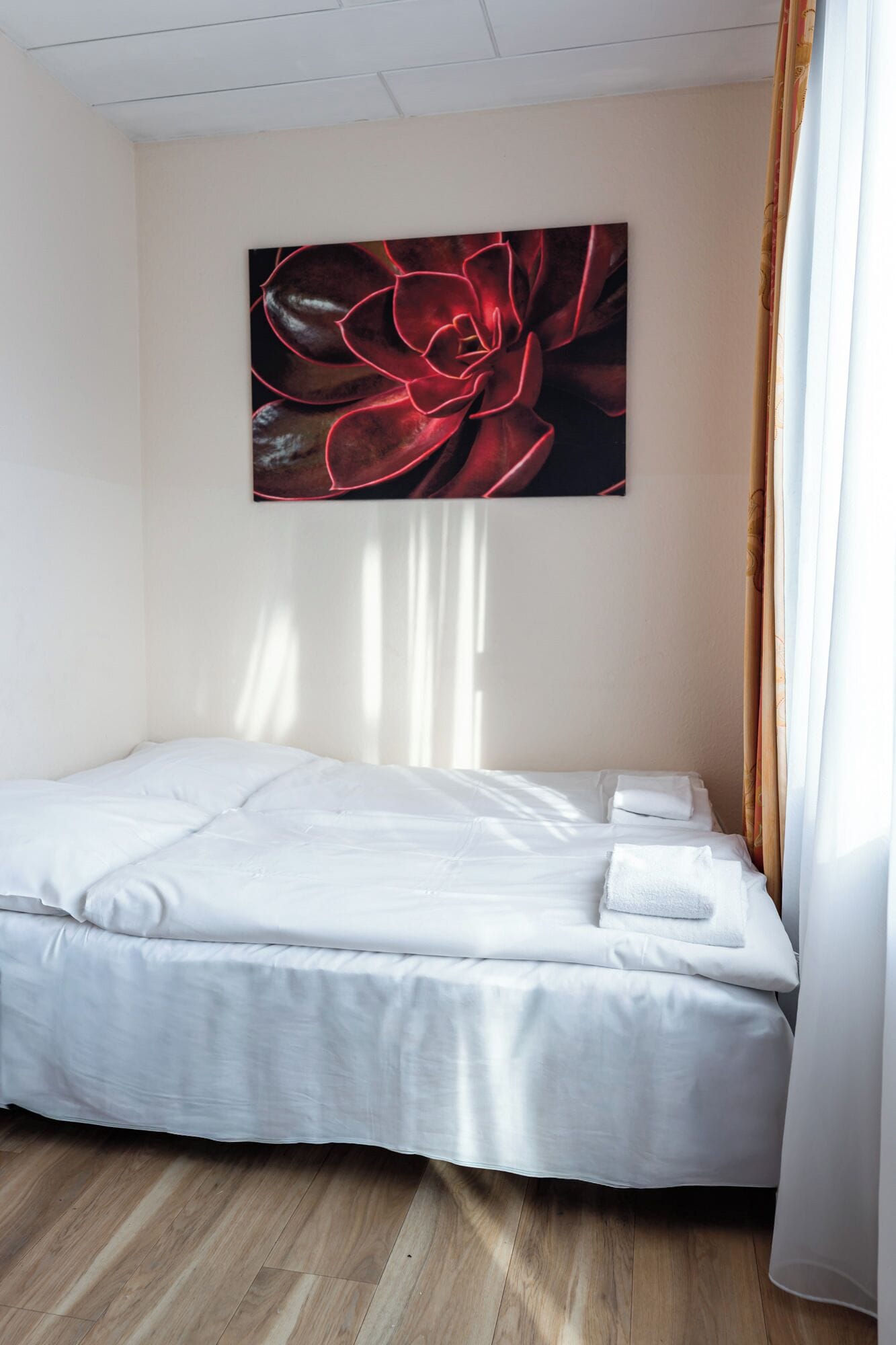 Una cama individual con sábanas blancas en una habitación con piso de madera y un cuadro grande con motivos florales en la pared.