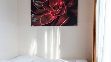 Una cama individual con sábanas blancas en una habitación con piso de madera y un cuadro grande con motivos florales en la pared.