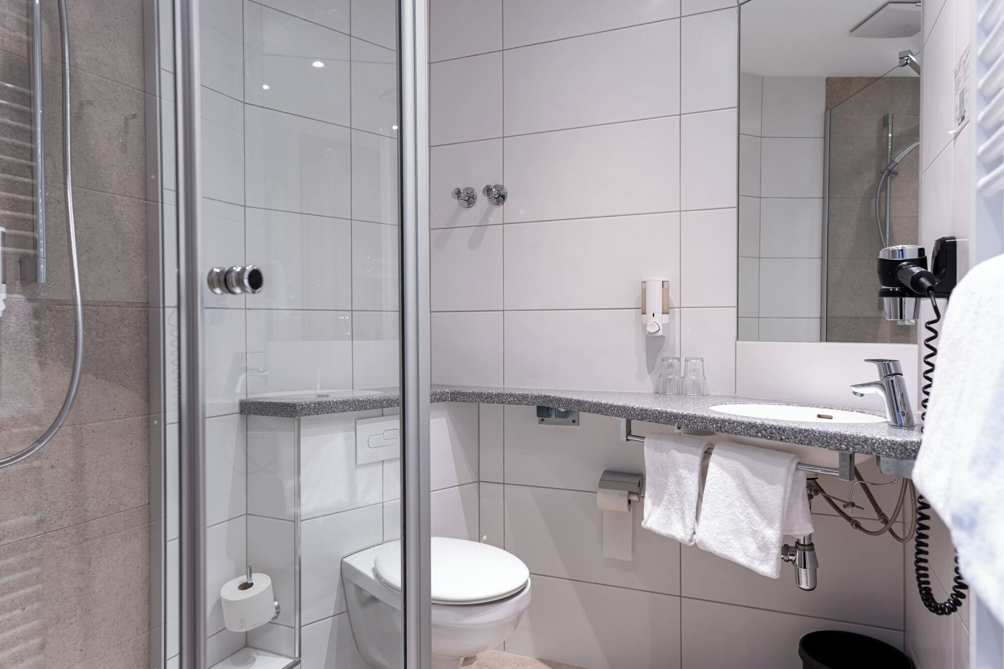 Un baño moderno con ducha de vidrio, inodoro, lavamanos con toallero y espejo.
