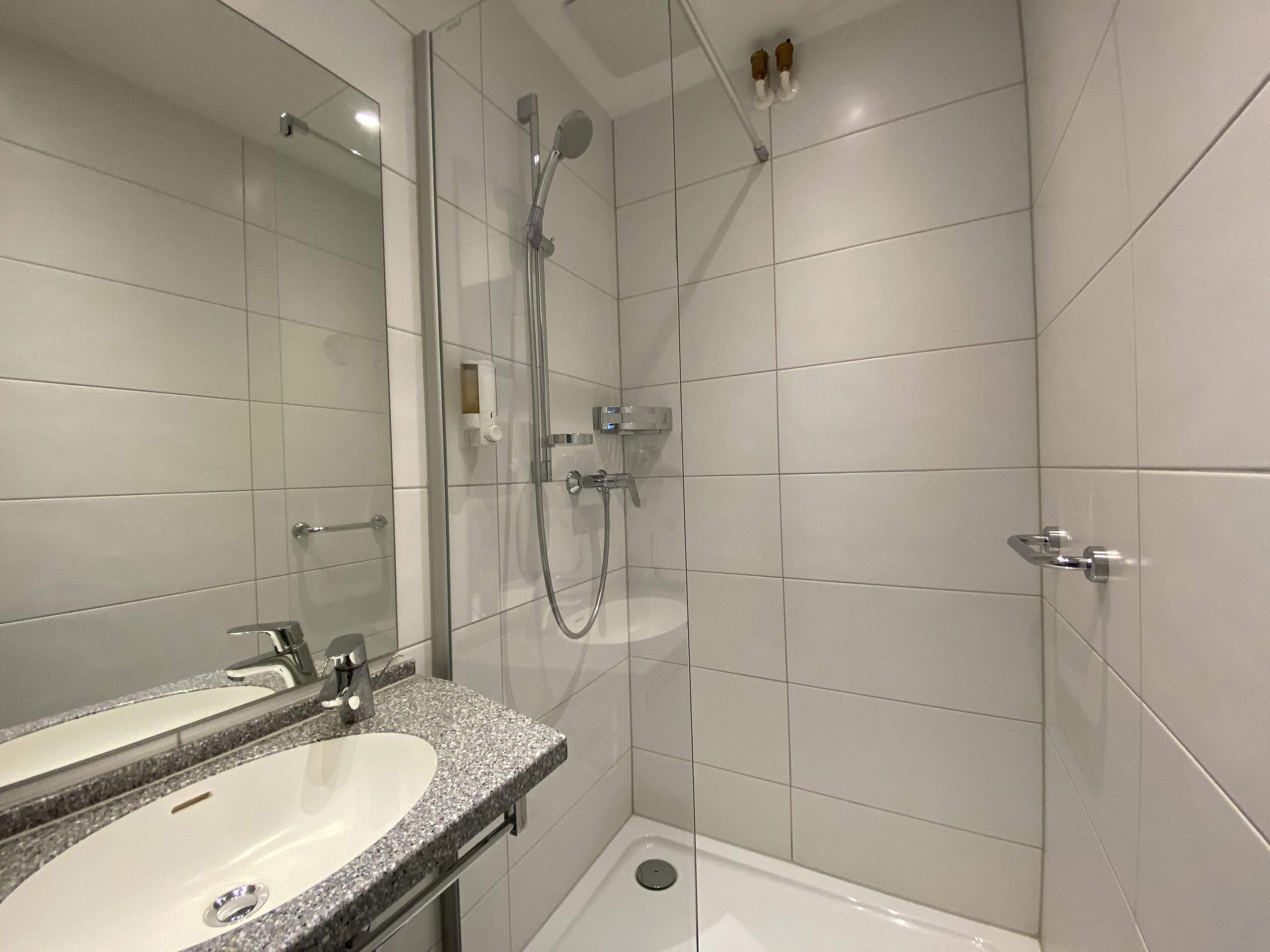 Un baño moderno con tocador de doble lavabo, cabina de ducha con mampara de vidrio y suelo de baldosas blancas.