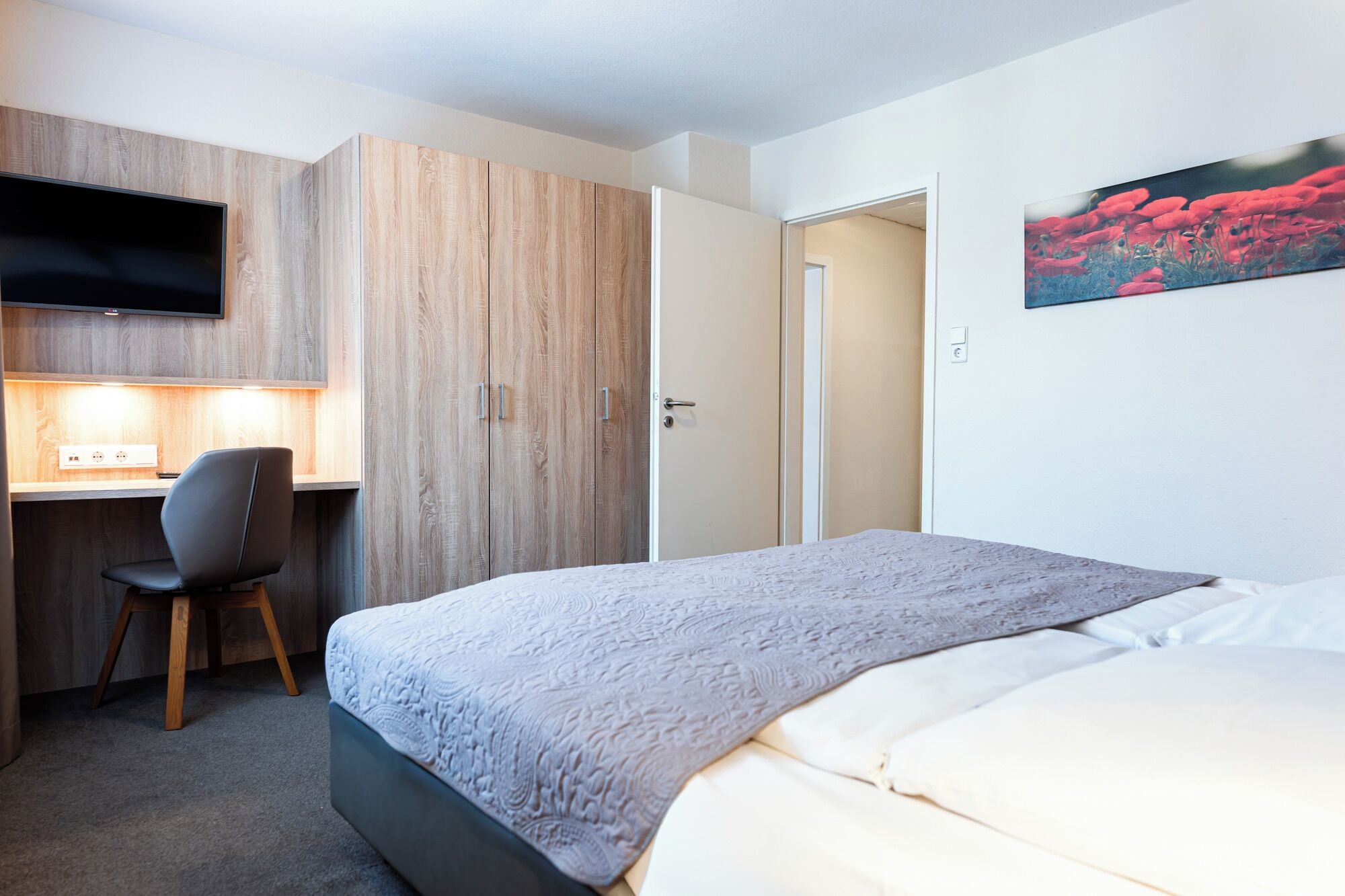 A hotel room with a bed, a desk with a chair, a television mounted on the wall, and a door leading to another room.