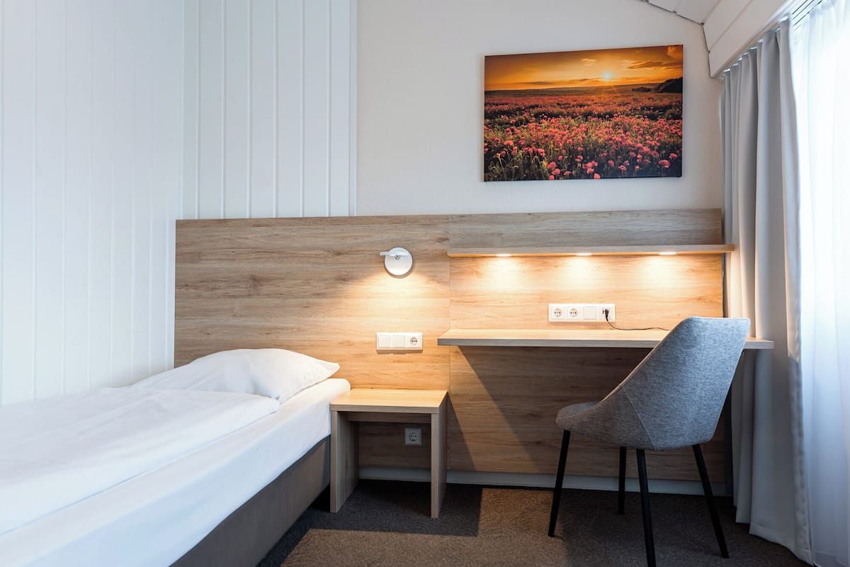 Una habitación pequeña con una cama, un escritorio con silla, una lámpara de pared y un cuadro de un campo.