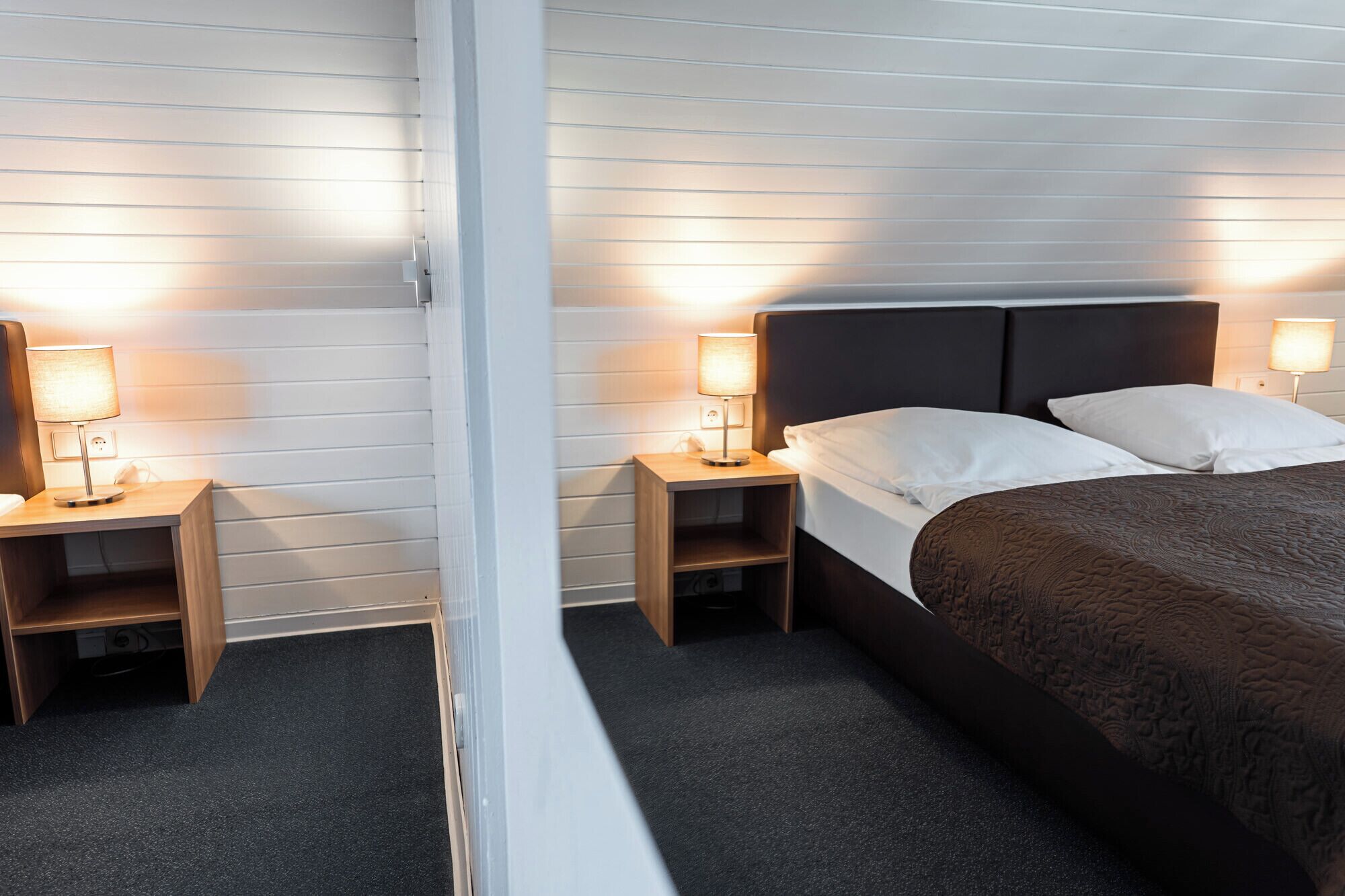 A hotel room with a bed, two bedside tables with lamps, a wooden headboard, and a carpeted floor.