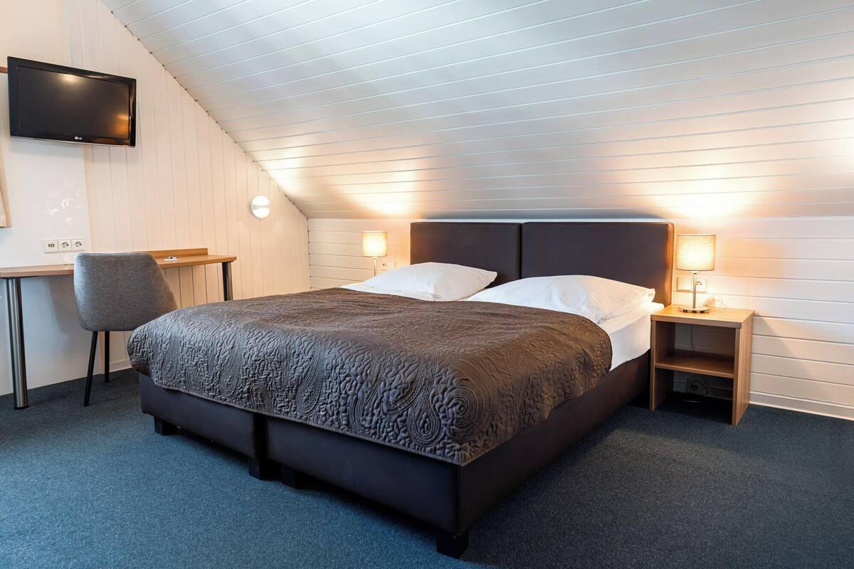 Una habitación de hotel moderna con una cama grande, dos lámparas de noche, un televisor montado en la pared y un escritorio con una silla.