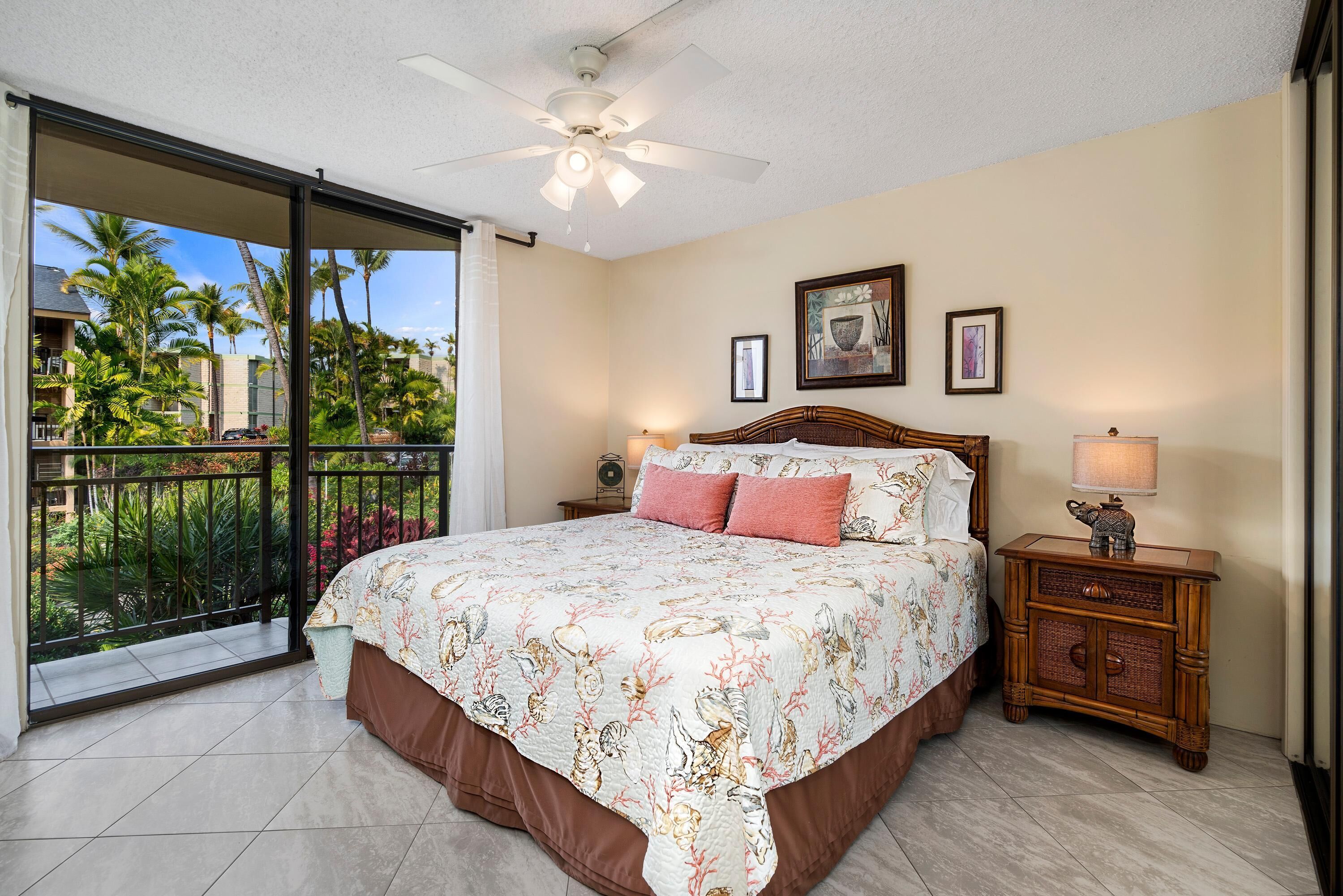 A bedroom with a large bed, a ceiling fan, a nightstand, and a view of palm trees through a sliding glass door.