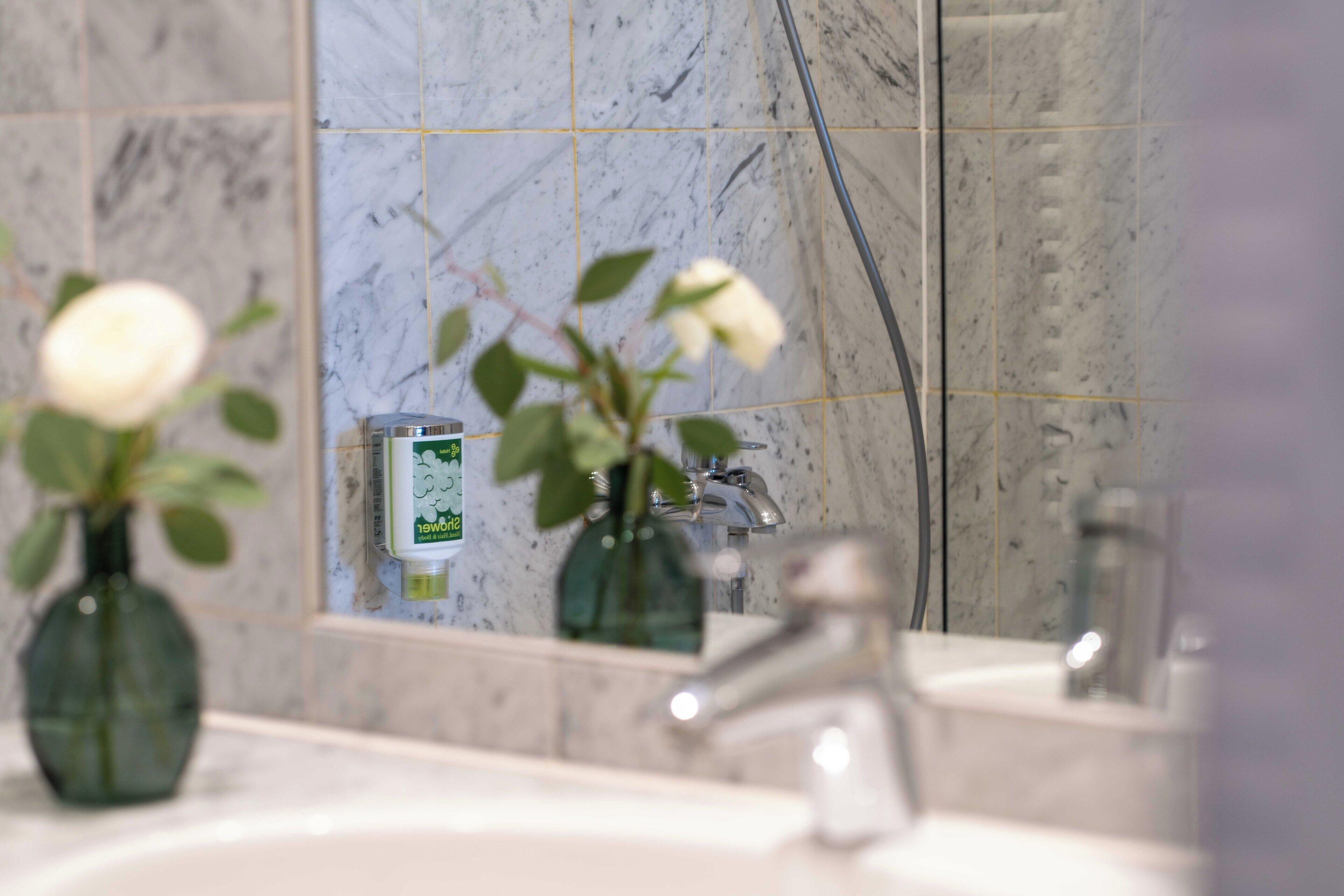 A bathroom with a marble wall, a mirror, a sink, and a green vase with white flowers.