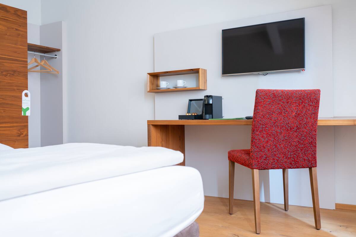 Une chambre d’hôtel comprenant un lit, un bureau avec une chaise, une télévision et une étagère avec des tasses.