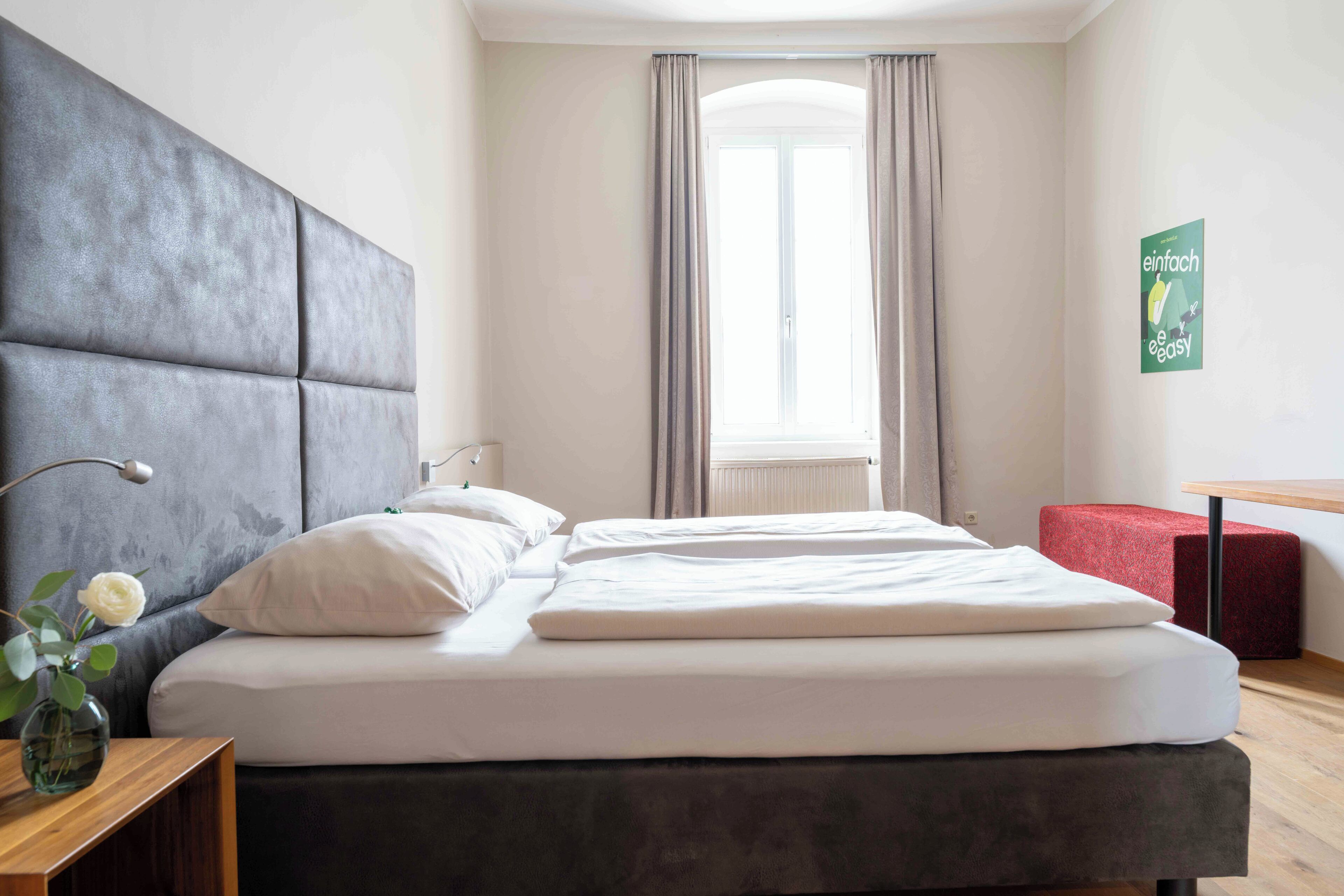 A modern hotel room with a large bed, a bedside table with a vase, and a window with curtains.