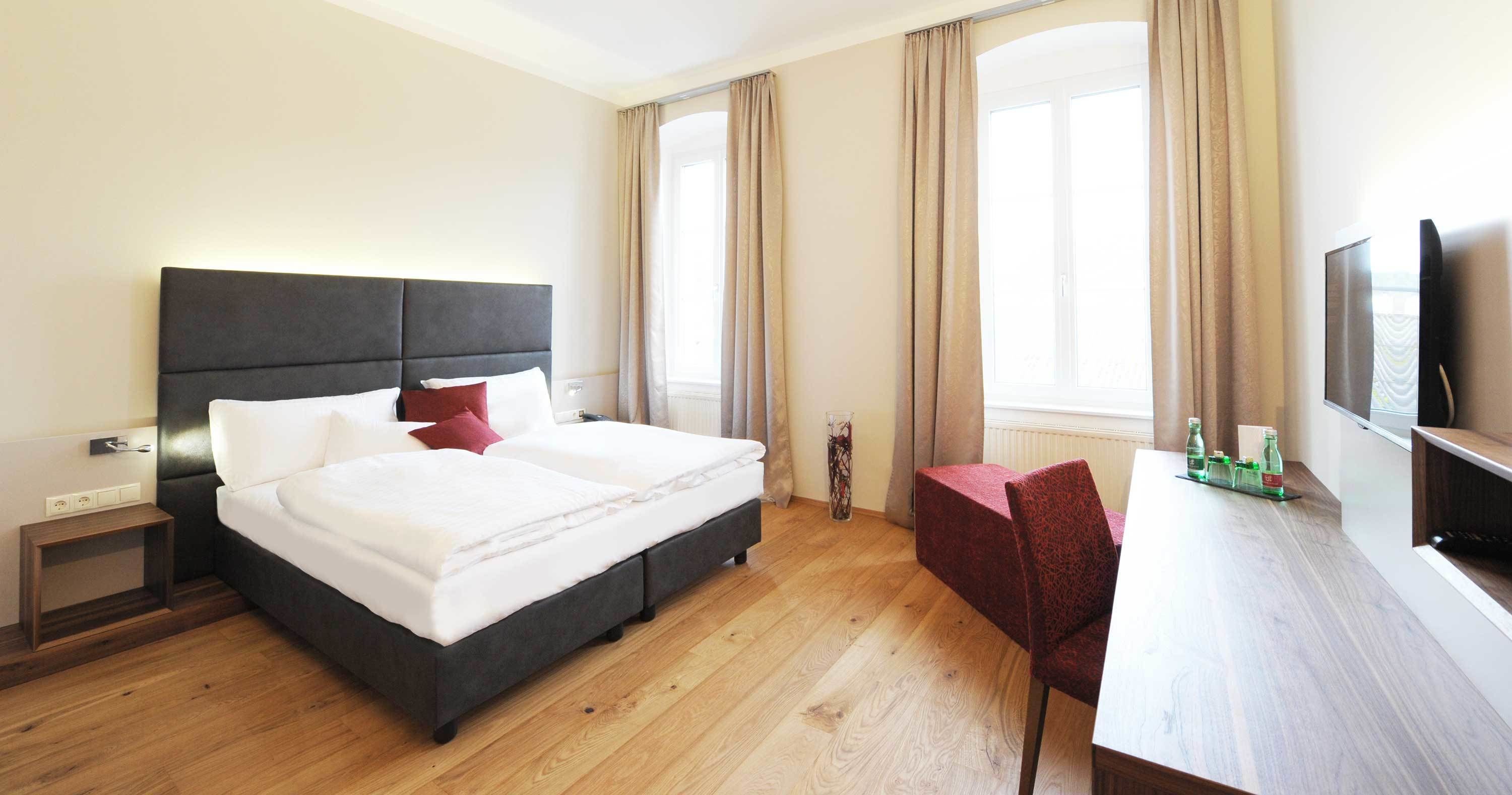 Habitación de hotel con cama, mesita de noche, silla, escritorio, televisor y ventanas amplias.