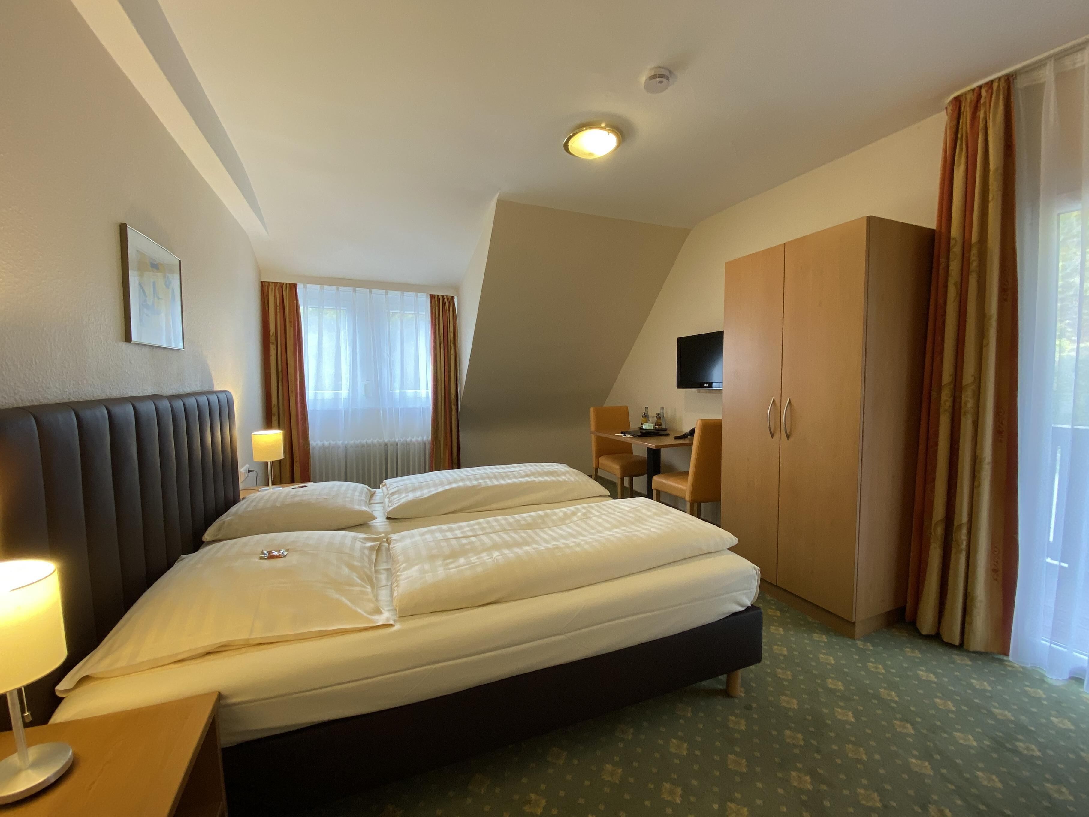 A hotel room with a large bed, a desk, a chair, a television, and a wardrobe.