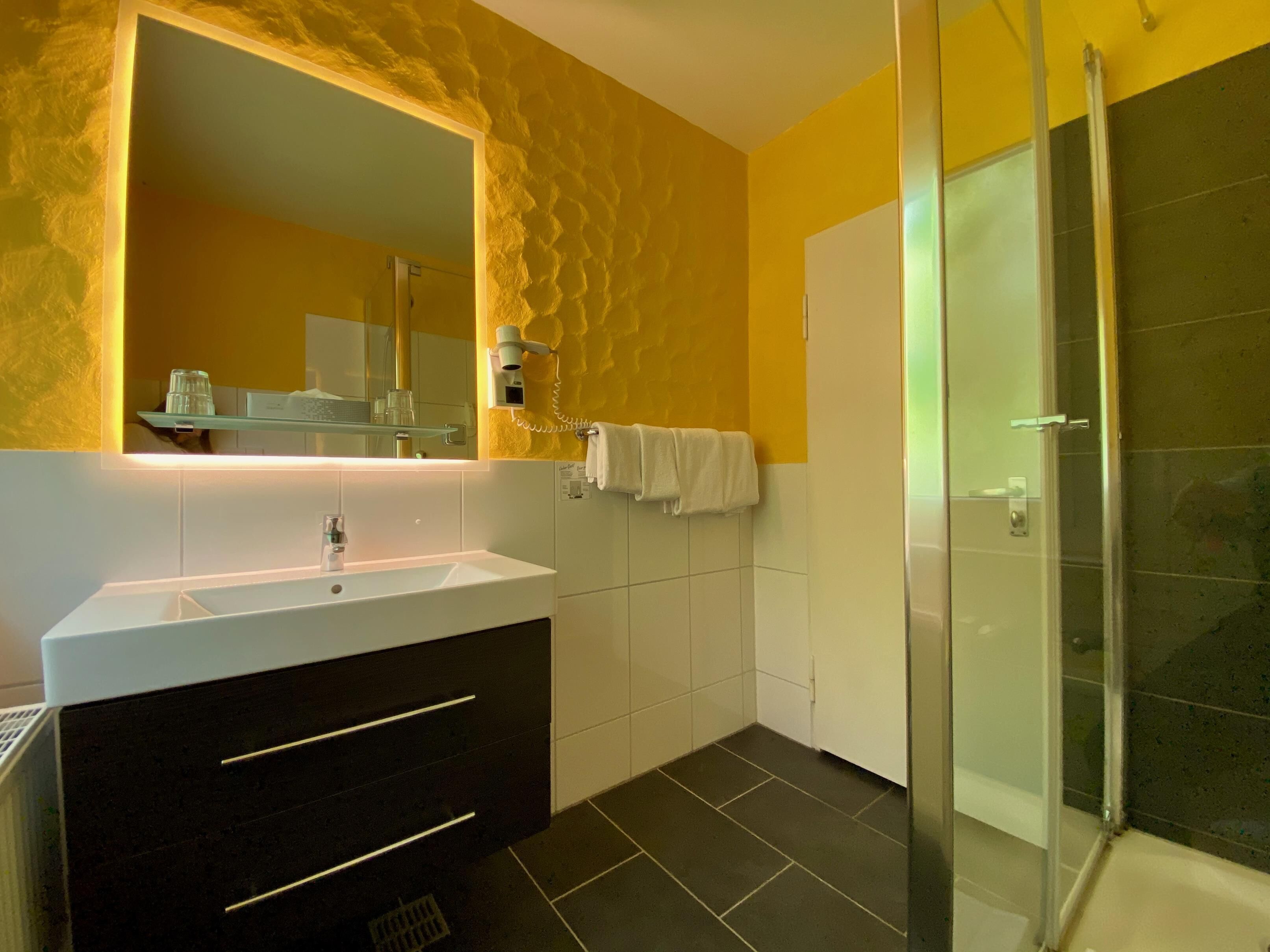 A modern bathroom with a large mirror, a white sink, a dark vanity, a glass shower enclosure, and a yellow textured wall.