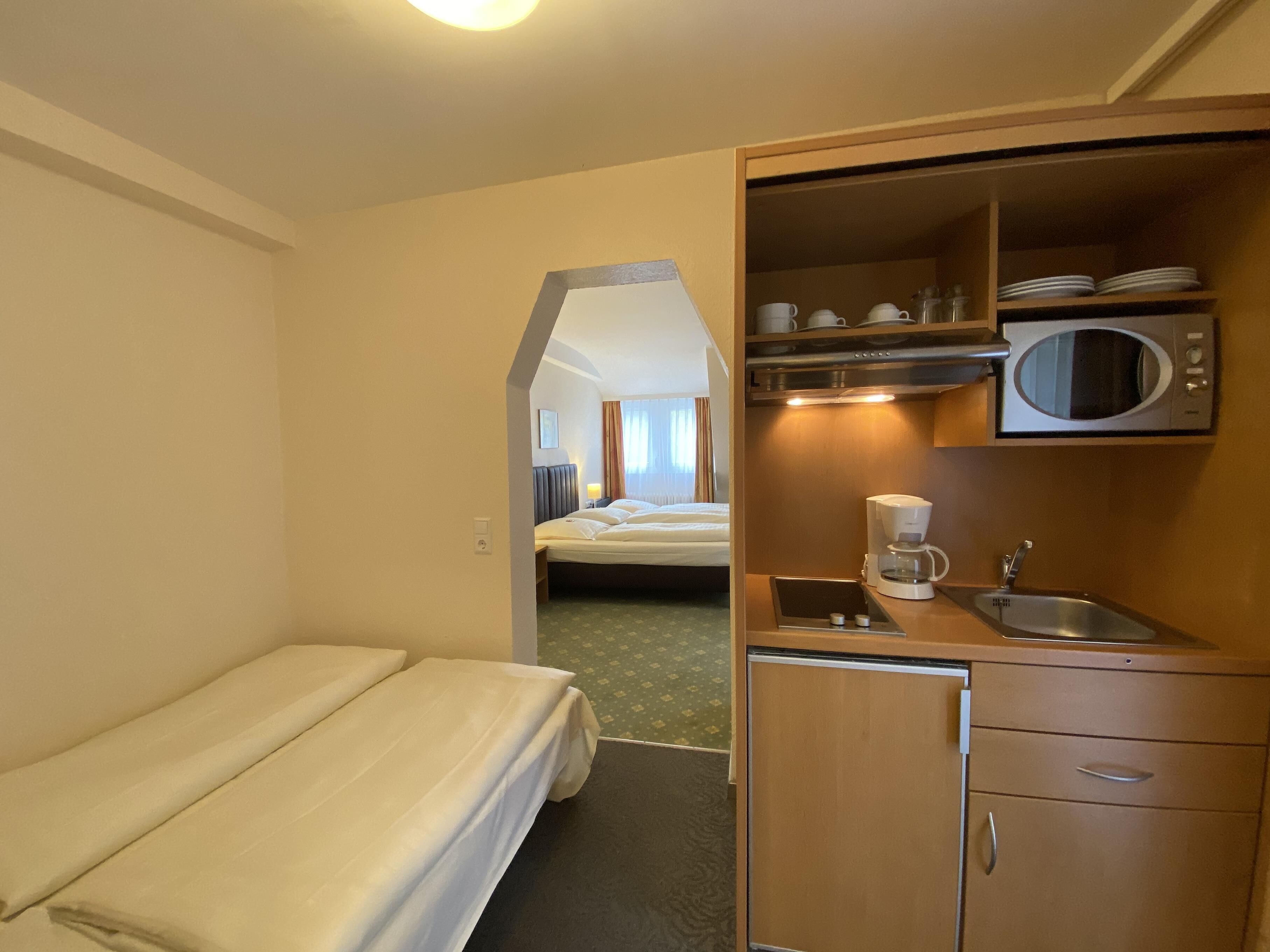 A compact hotel room with a single bed, a kitchenette with a microwave, coffee maker, and sink, and a small bathroom visible through an archway.