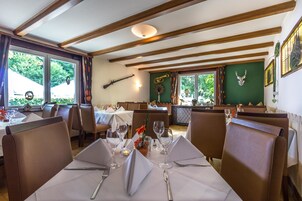 A restaurant with wooden beams on the ceiling, tables set with white tablecloths, and various wall decorations including a wreath and a musical instrument.