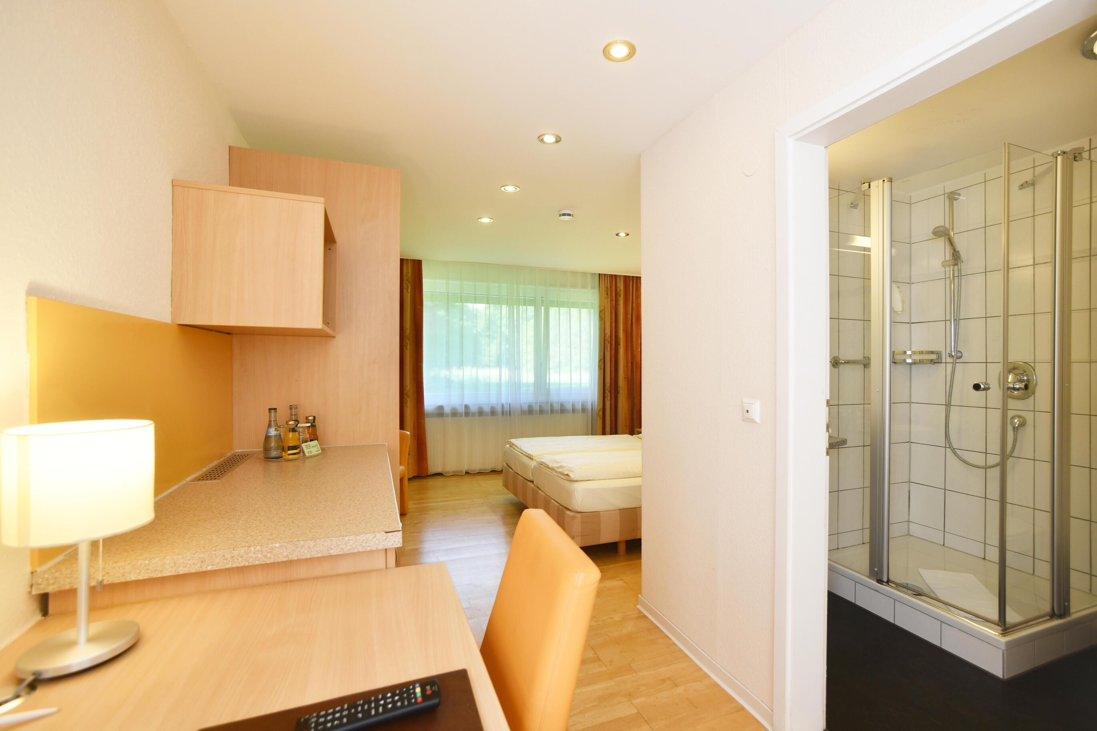 A hotel room with a desk, chair, bed, and a glass-enclosed shower area.