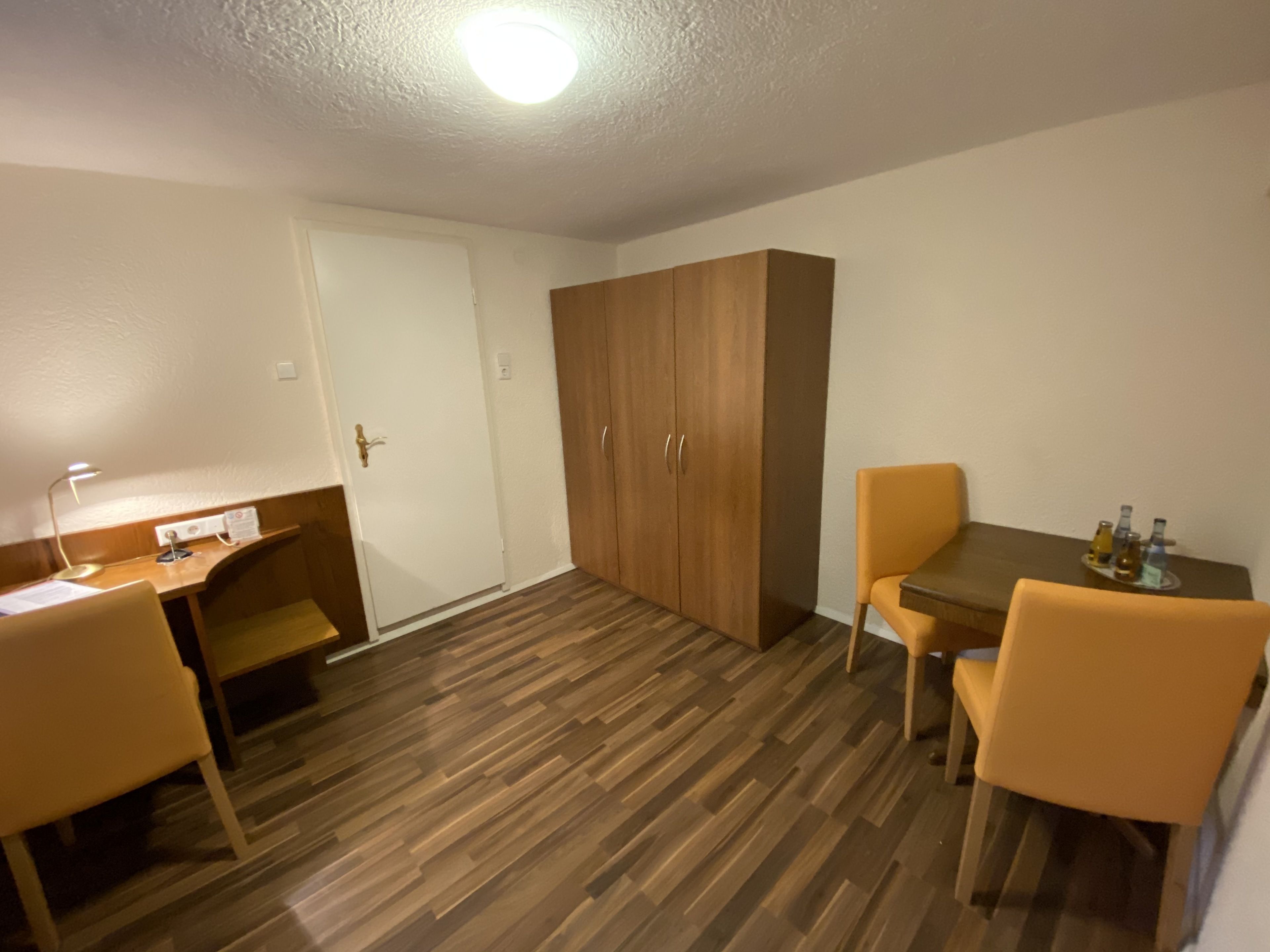 Una habitación con suelo de madera, un escritorio con lámpara, un armario y una mesa de comedor con sillas.