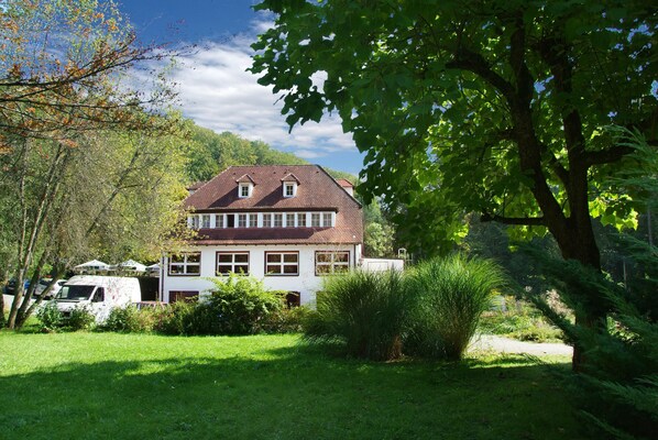 Une maison à deux étages, dotée d’un toit en pente, entourée d’un jardin bien entretenu et d’arbres.