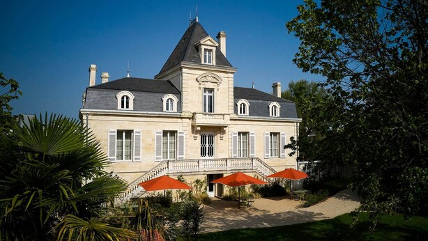 A large, two-story building with a prominent tower, a balcony with red umbrellas, and a well-maintained garden.
