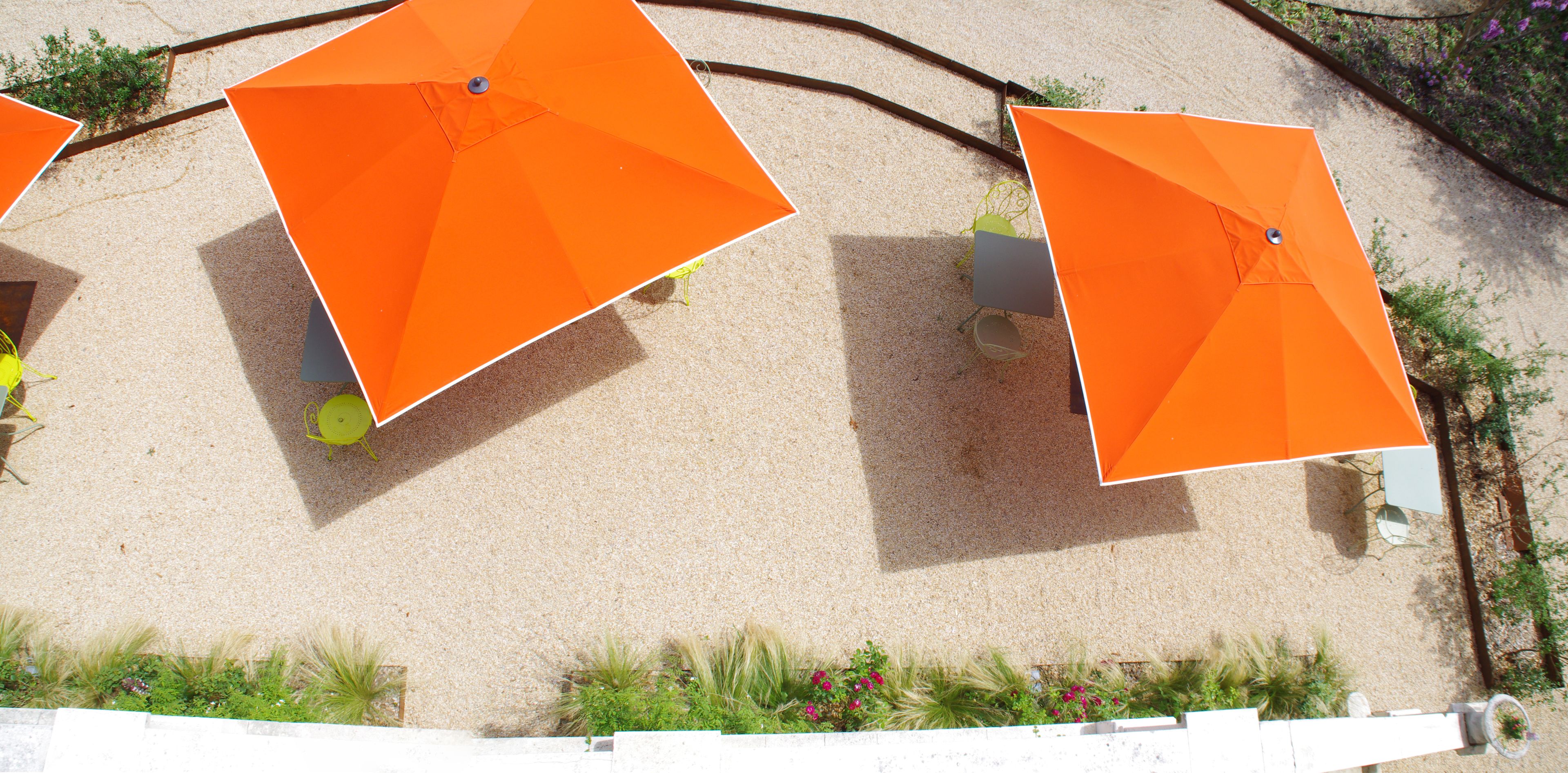 Blick von oben auf eine Sandfläche mit orangefarbenen Sonnenschirmen und Sitzgelegenheiten.