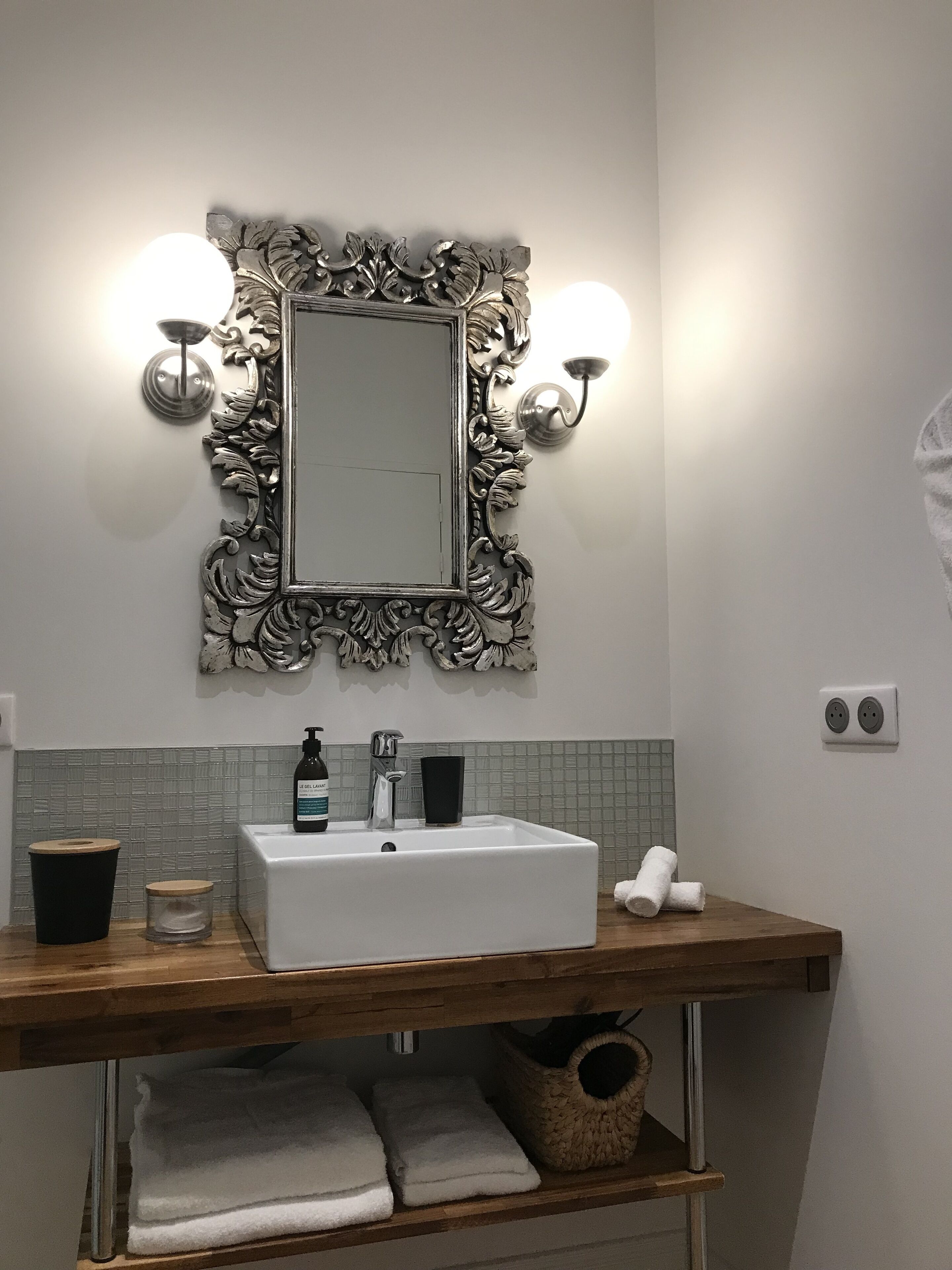 Une salle de bain avec un lavabo blanc, un miroir doté d’un cadre argenté orné, deux appliques murales, un distributeur de savon et un porte-serviettes avec des serviettes.