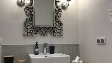 Une salle de bain avec un lavabo blanc, un miroir doté d’un cadre argenté orné, deux appliques murales, un distributeur de savon et un porte-serviettes avec des serviettes.