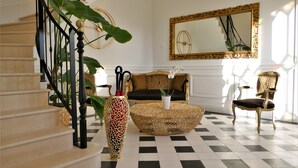 A staircase with a black balustrade, a round coffee table with a vase, a sofa with patterned cushions, and a large ornate mirror on the wall.