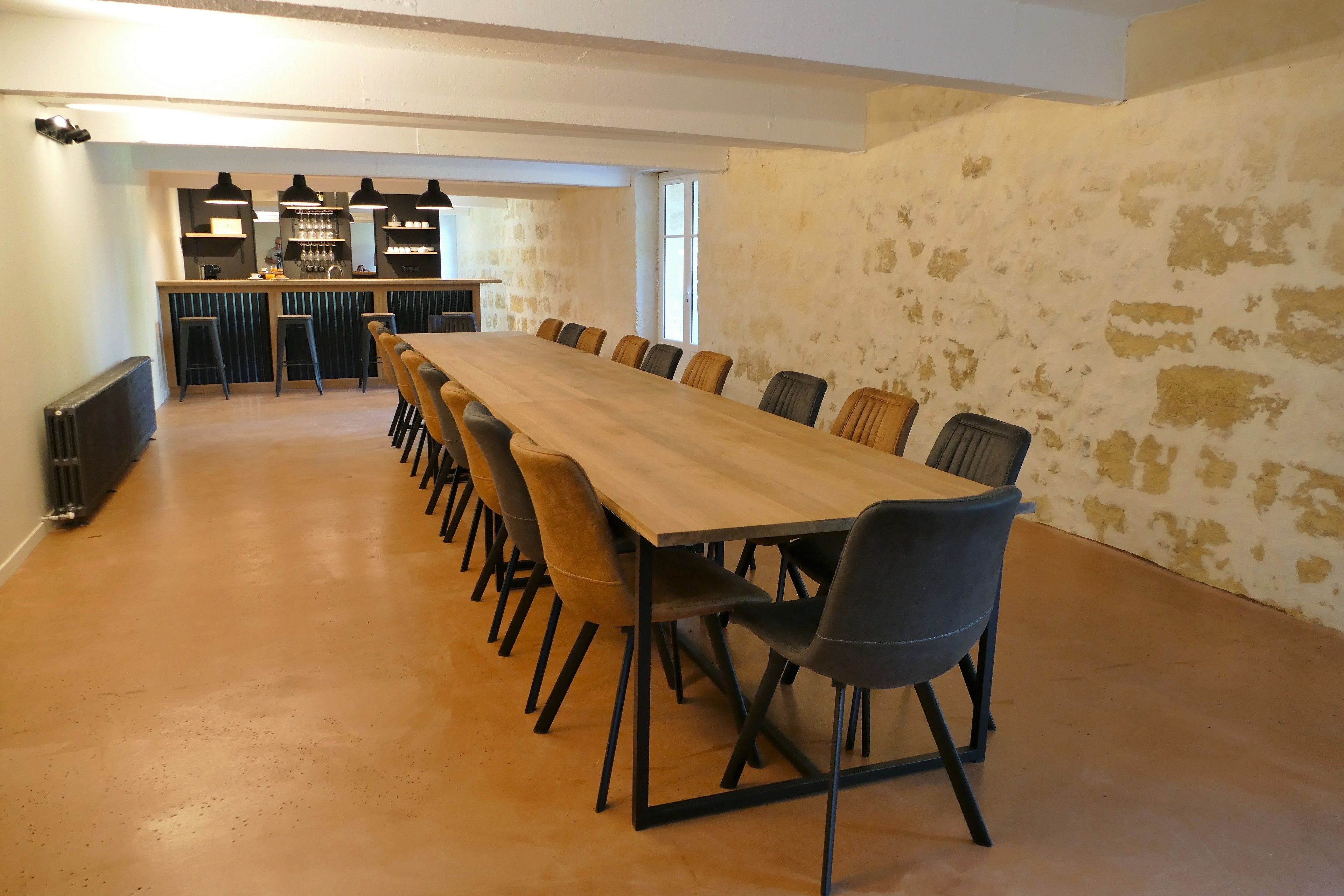 A long wooden table with chairs, a kitchen area with bar stools, and a stone wall.
