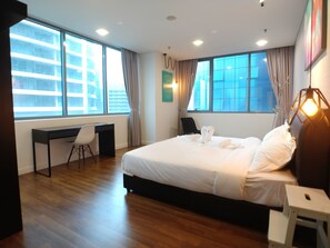 A modern hotel room with a large bed, a desk, and a chair. There are two windows with curtains, and a wall-mounted light fixture. - OHO Suites KLCC Summer Suites (Kuala Lumpur)