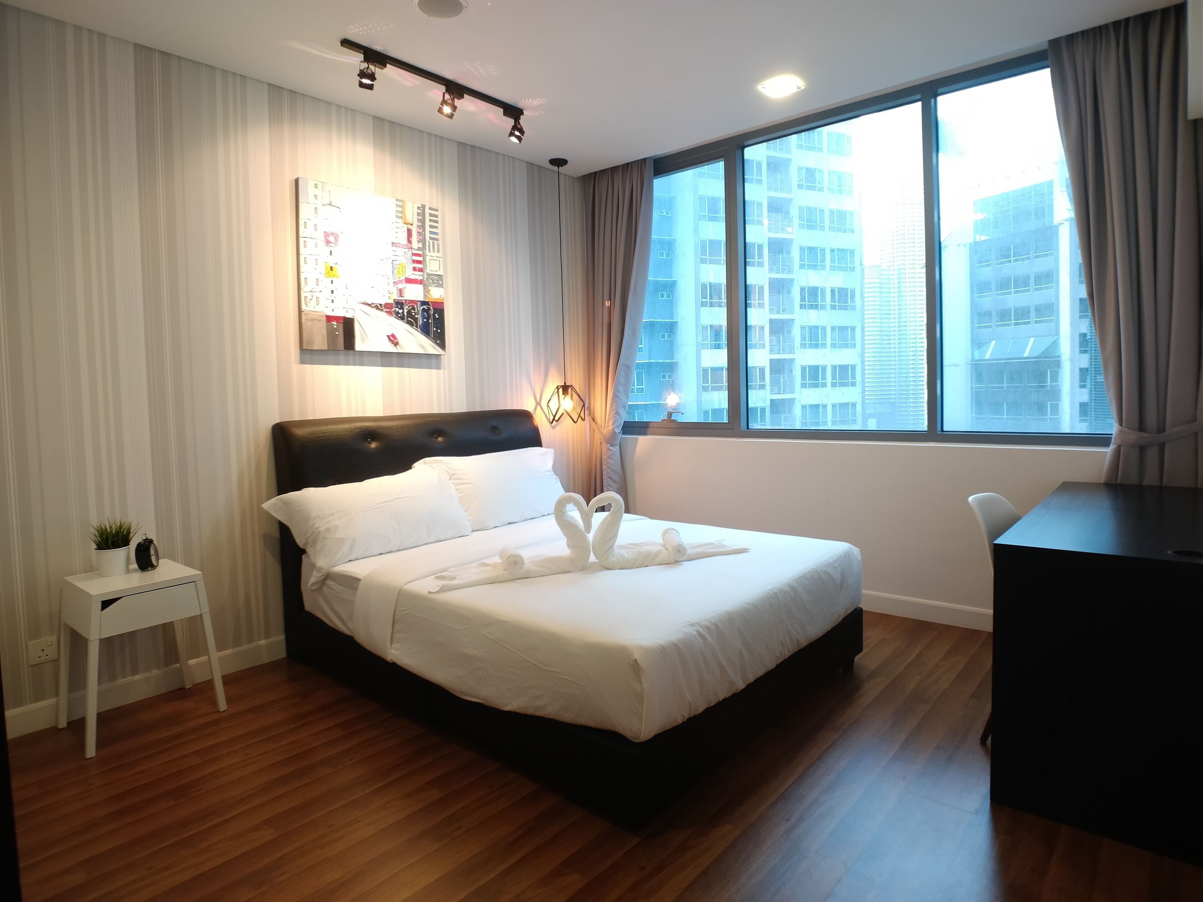 A modern hotel room with a large bed, a small bedside table with a plant, a desk, and a large window with a city view.