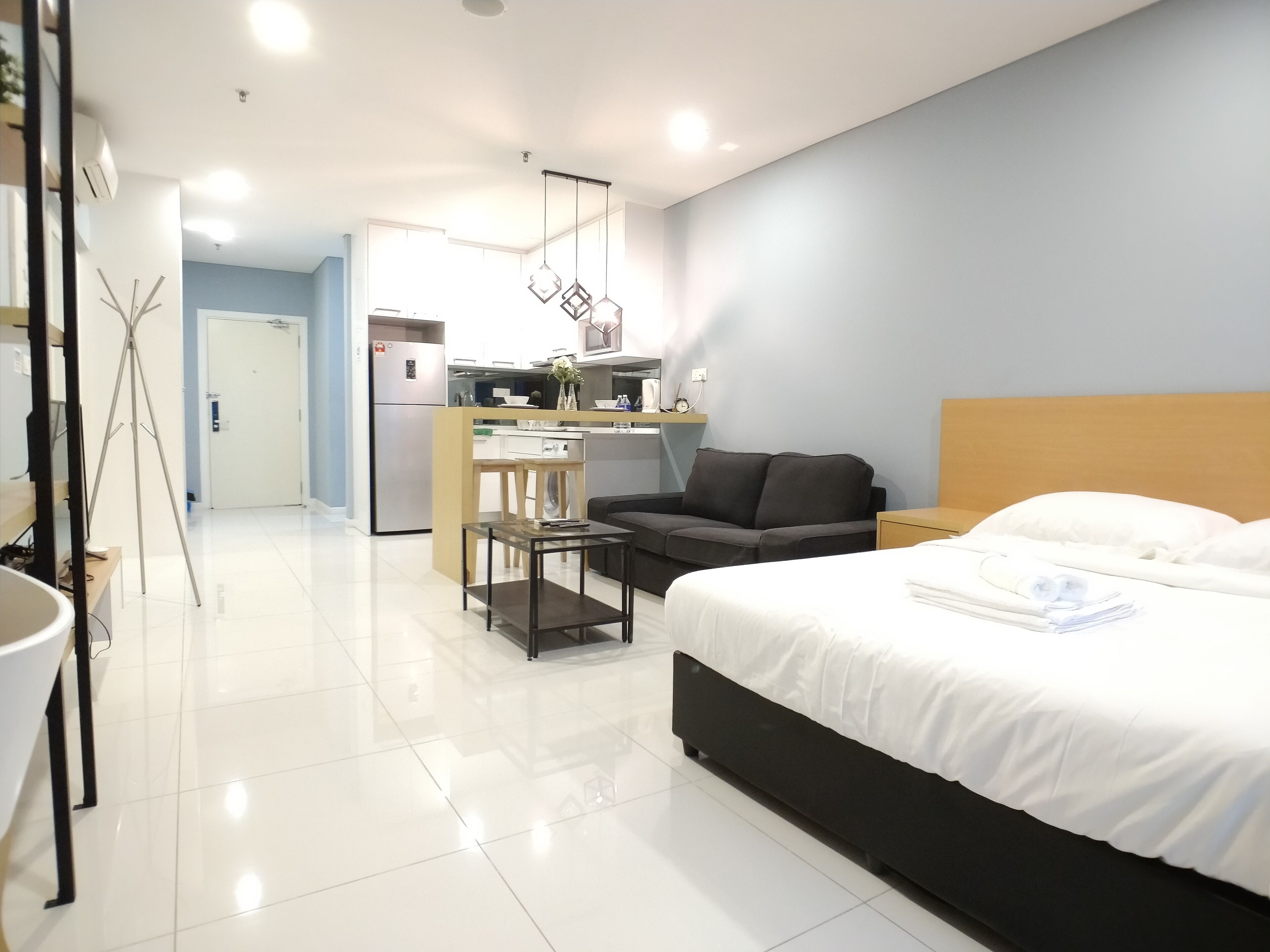 A modern hotel room with a large bed, a small sofa, a desk, and a kitchenette in the background.