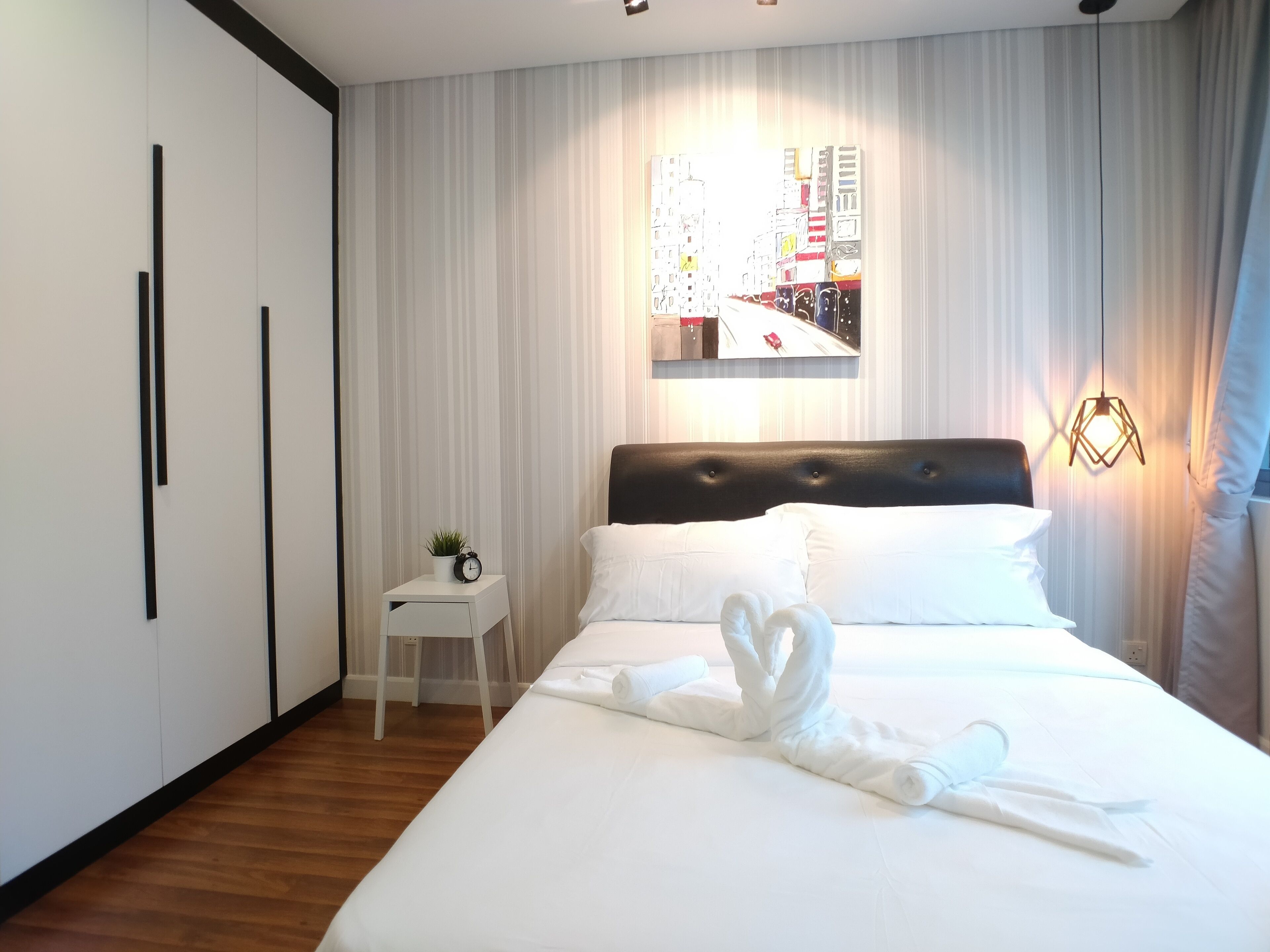 A hotel room with a neatly made bed, a small bedside table with a clock and a plant, a large window with curtains, and a wall-mounted artwork.