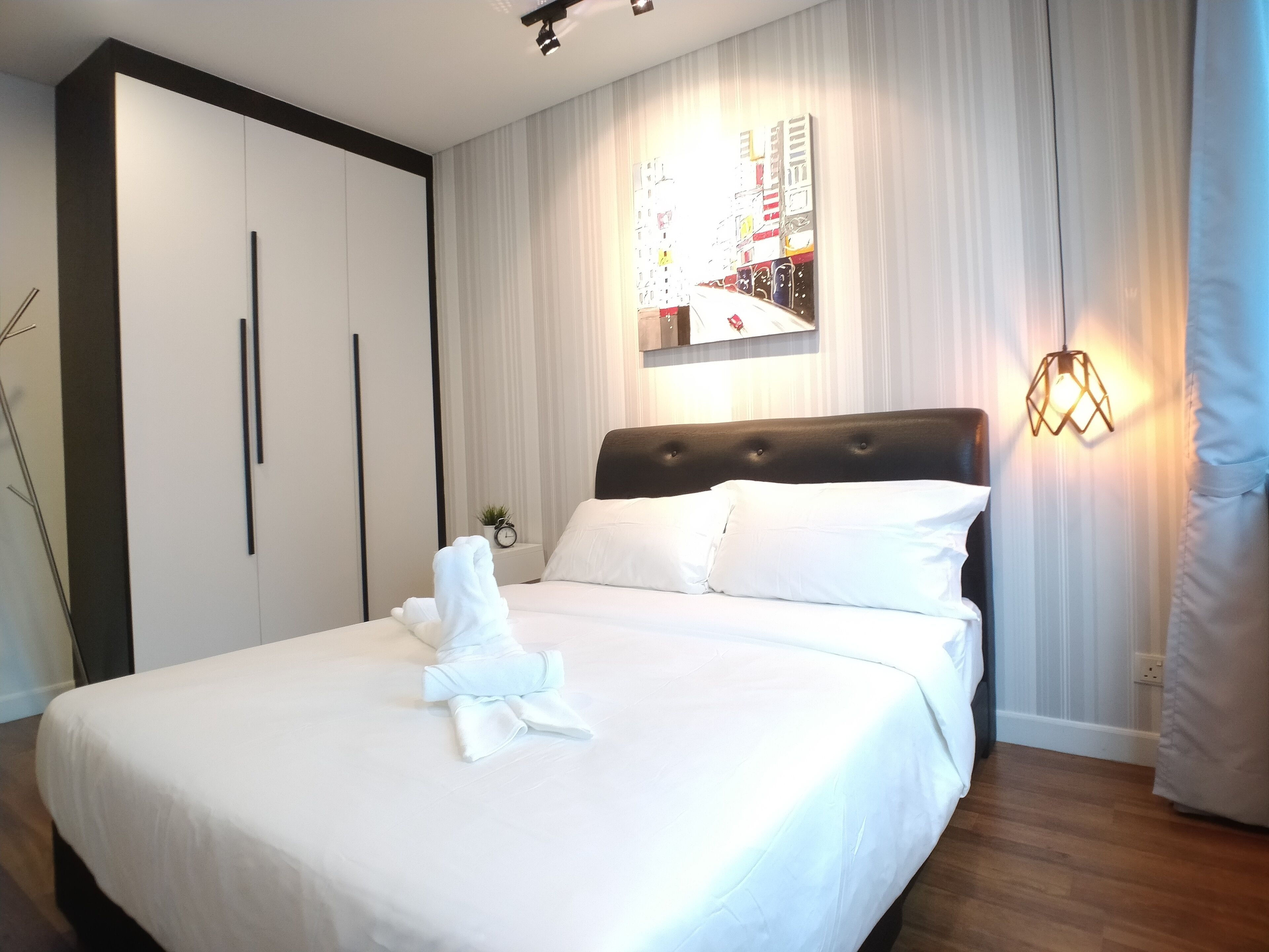 A neatly made bed with white linens and a folded towel, a black headboard, and a wall-mounted light fixture.