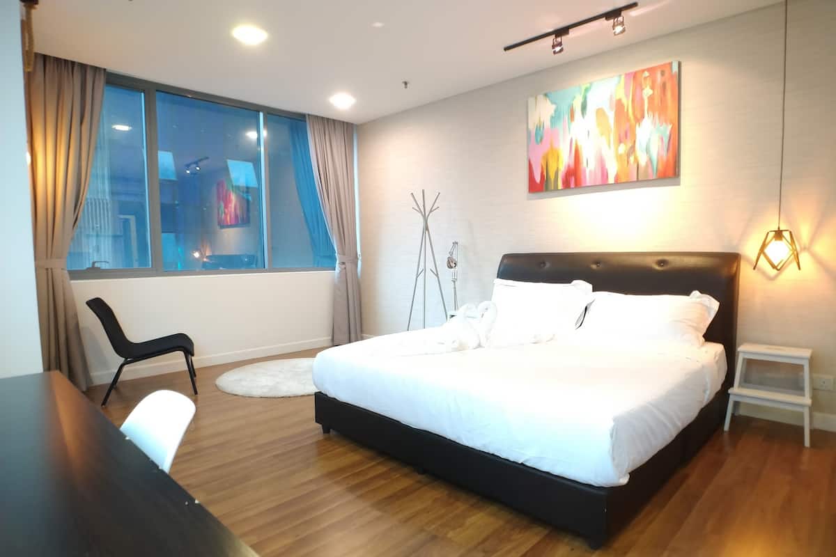 Une chambre d’hôtel moderne avec un grand lit, un bureau, une chaise, une fenêtre avec des rideaux, une lampe sur pied et une peinture abstraite aux couleurs vives accrochée au mur.