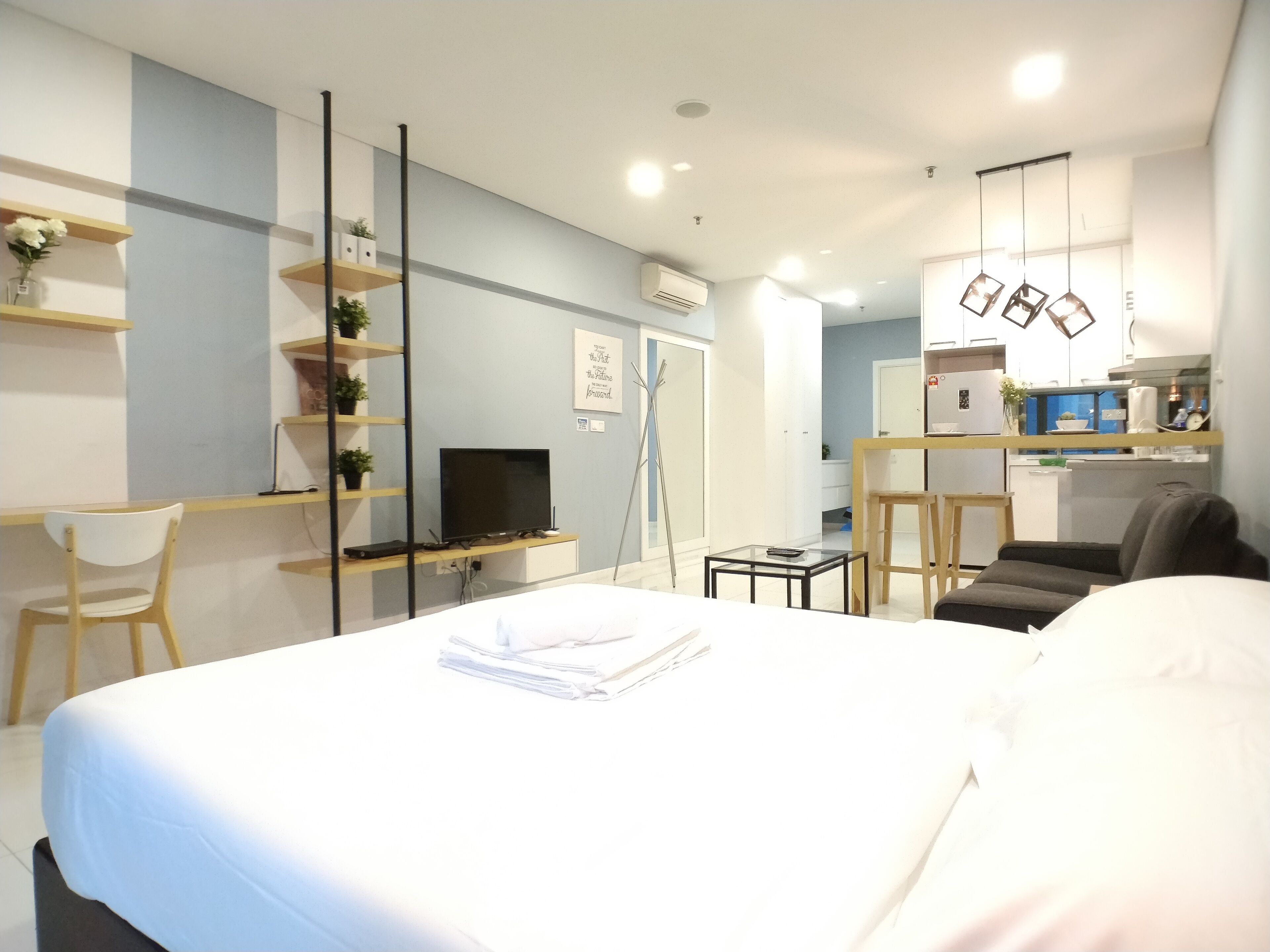 Ein modernes Hotelzimmer mit einem großen Bett, einem Schreibtisch mit Computer, einem Sessel und einem kleinen Tisch. Im Hintergrund ist eine Küchenzeile zu sehen.