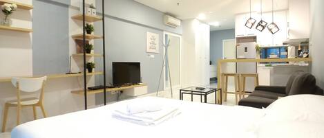Habitación de hotel moderna con una cama grande, un escritorio con una computadora, una silla y una mesita. Se observa una kitchenette al fondo.