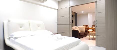 Un dormitorio moderno con una cama grande, un armario empotrado y un área de comedor visible a través de una puerta abierta.