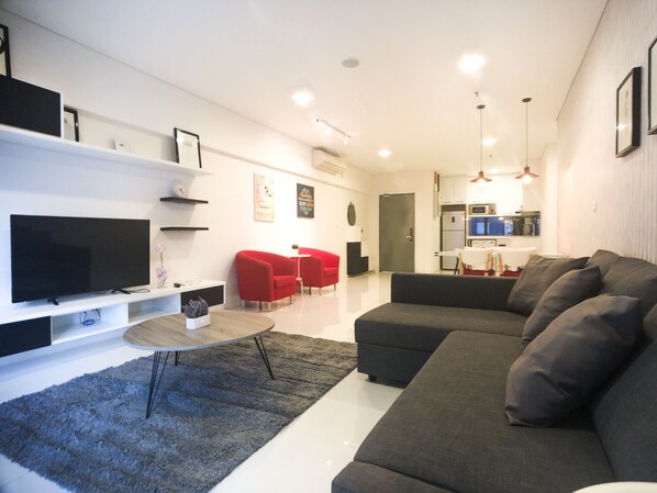A modern living room with a large flat-screen TV, a grey sofa, red chairs, and a kitchen in the background. - OHO Suites KLCC Summer Suites (Kuala Lumpur)