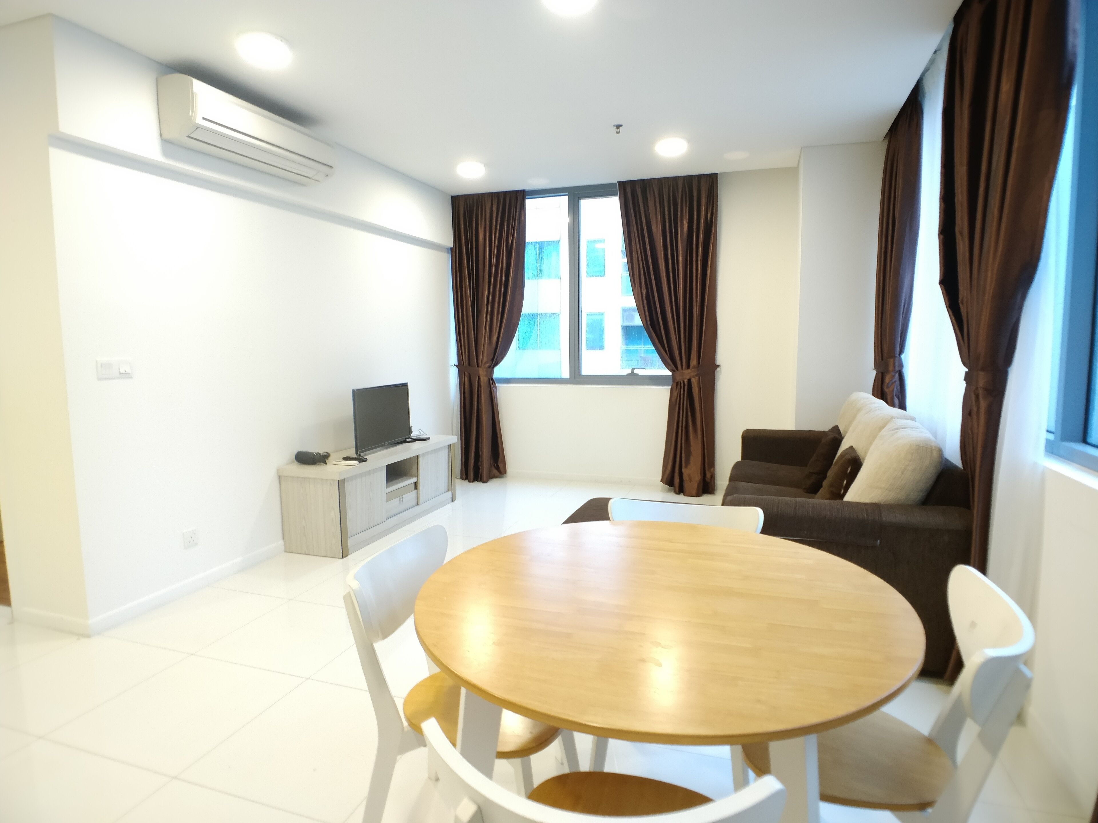 A modern living room with a round wooden table, white chairs, a grey sofa, and a TV on a wooden stand.