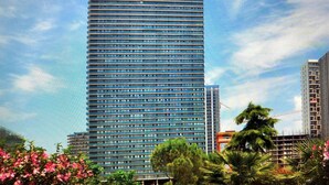 A tall, modern skyscraper with a grid of windows, surrounded by lush greenery and flowering plants. - Hotel 36 - ORBI Beach Tower Batumi 36th floor (Batumi)